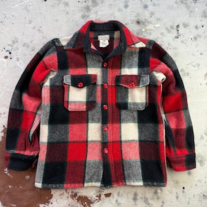 Op de afbeelding: Een vintage rood, zwart en wit geruit wollen overhemd met twee borstzakken en rode knopen. Het overhemd heeft een klassieke kraag en lange mouwen. De stof lijkt dik en warm.