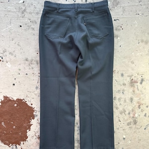 Puede incluir: Pantalones grises oscuros con dos bolsillos traseros y una pequeña etiqueta. Los pantalones tienen un diseño de pierna recta y están hechos de una tela suave. Los pantalones están colocados sobre una superficie desgastada.