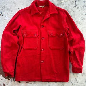 May include: A vibrant red wool shirt jacket featuring two chest pockets and button fastenings. The jacket has a traditional collar and buttoned cuffs. The garment is displayed on a distressed, light-coloured surface.