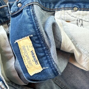 May include: Close-up of blue denim jeans with a Wrangler label. The label indicates a size of 36x32. The jeans have a classic five-pocket design and a button and zipper closure. The denim has a faded, worn appearance.