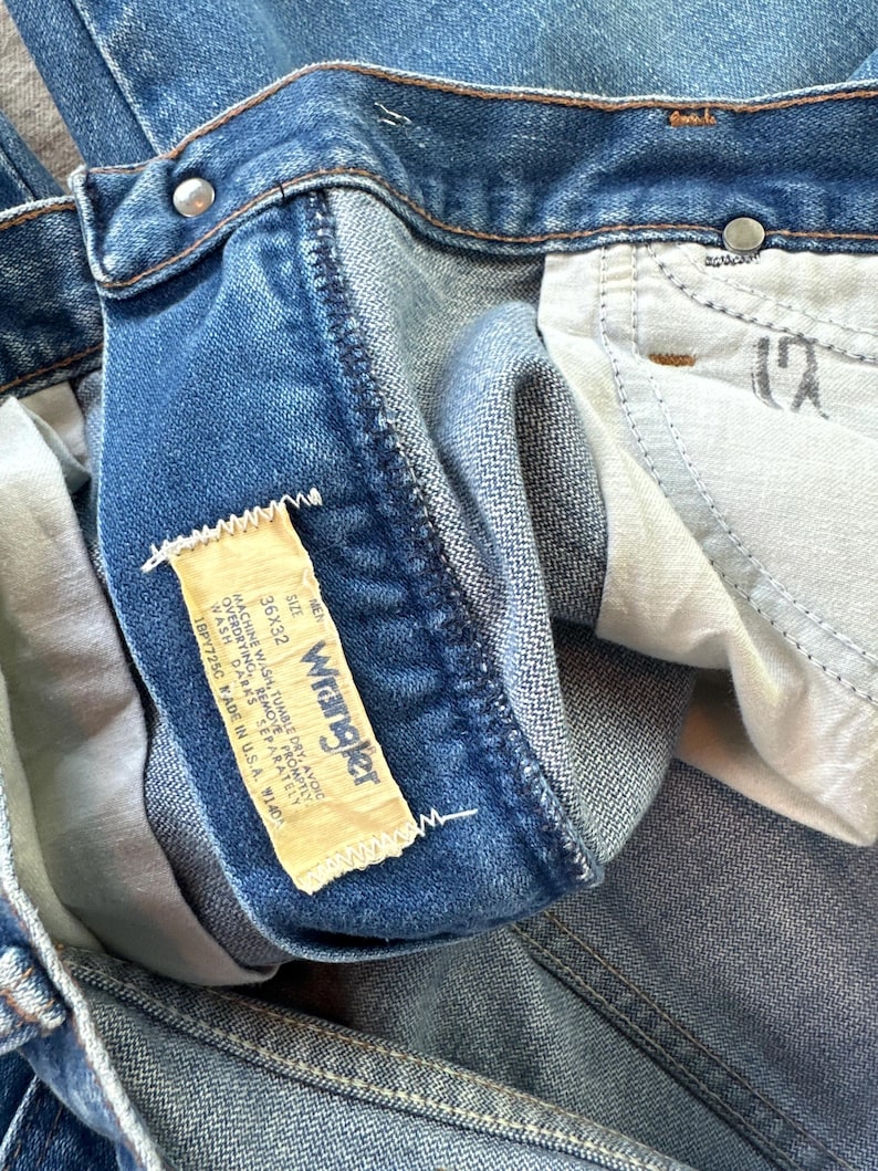 May include: Close-up of blue denim jeans with a Wrangler label. The label indicates a size of 36x32. The jeans have a classic five-pocket design and a button and zipper closure. The denim has a faded, worn appearance.