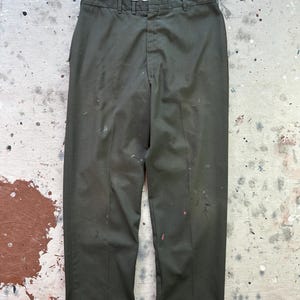 May include: Dark olive green trousers with a straight leg, featuring a button and zipper closure. The pants have belt loops and a white interior waistband. There are paint splatters on the front of the pants.