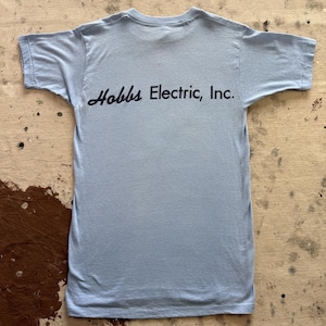 May include: A light blue short-sleeved t-shirt featuring the text Hobbs Electric, Inc. printed in black ink across the chest area. The garment has a crew neckline and a standard straight hemline, constructed from a solid-coloured cotton fabric.