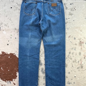 May include: Blue denim jeans with a classic five-pocket design. The jeans feature a straight leg cut and a brown leather patch on the back. The back pockets have a distinctive stitched design. The jeans are displayed on a distressed surface.