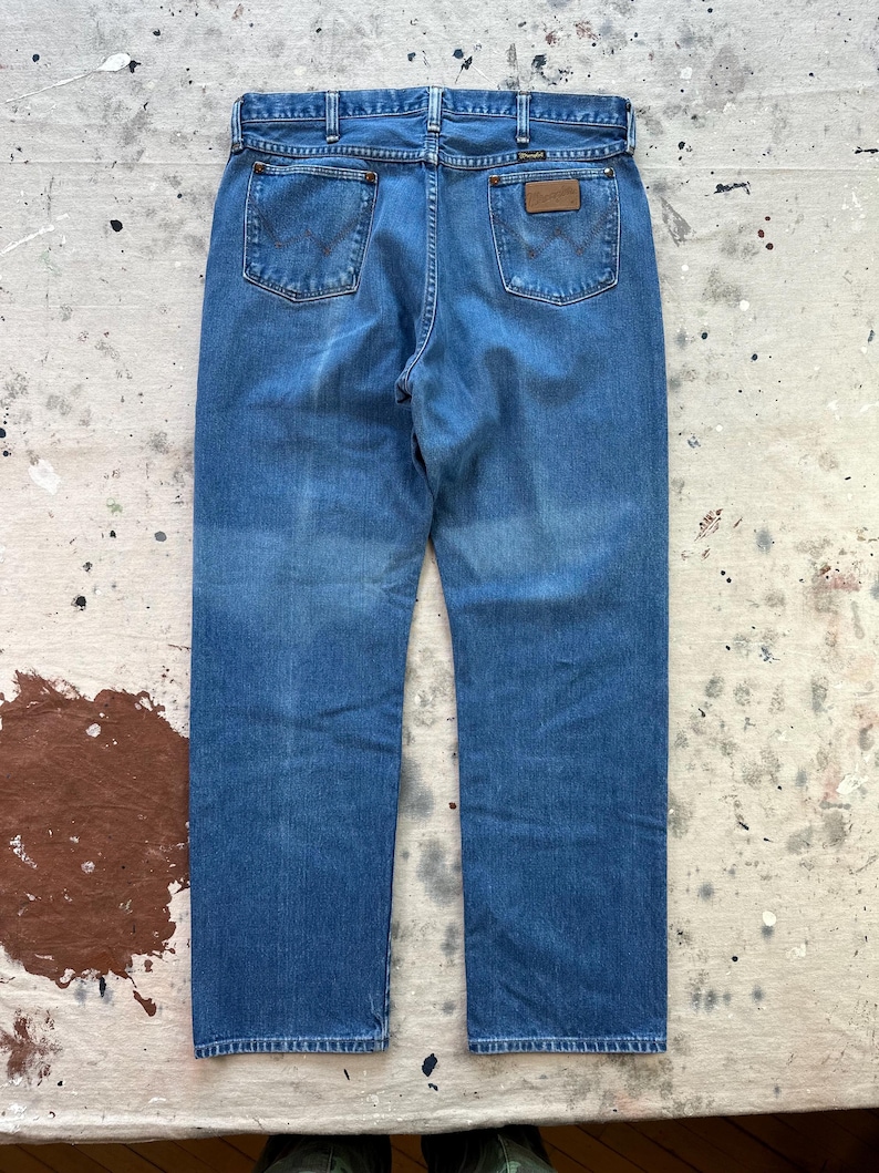 May include: Blue denim jeans with a classic five-pocket design. The jeans feature a straight leg cut and a brown leather patch on the back. The back pockets have a distinctive stitched design. The jeans are displayed on a distressed surface.