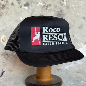 May include: Black trucker hat with a mesh back and a curved peak. The front displays the text "Roco RESCUE BATON ROUGE, LA" in white, with a red square graphic of a person abseiling. The hat is on a wooden stand.