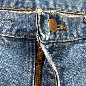 May include: Close-up of a pair of blue denim jeans. The image highlights the button and zipper area, with a brass-colored button and a zipper labeled "SCOVILL". The denim has a faded appearance.