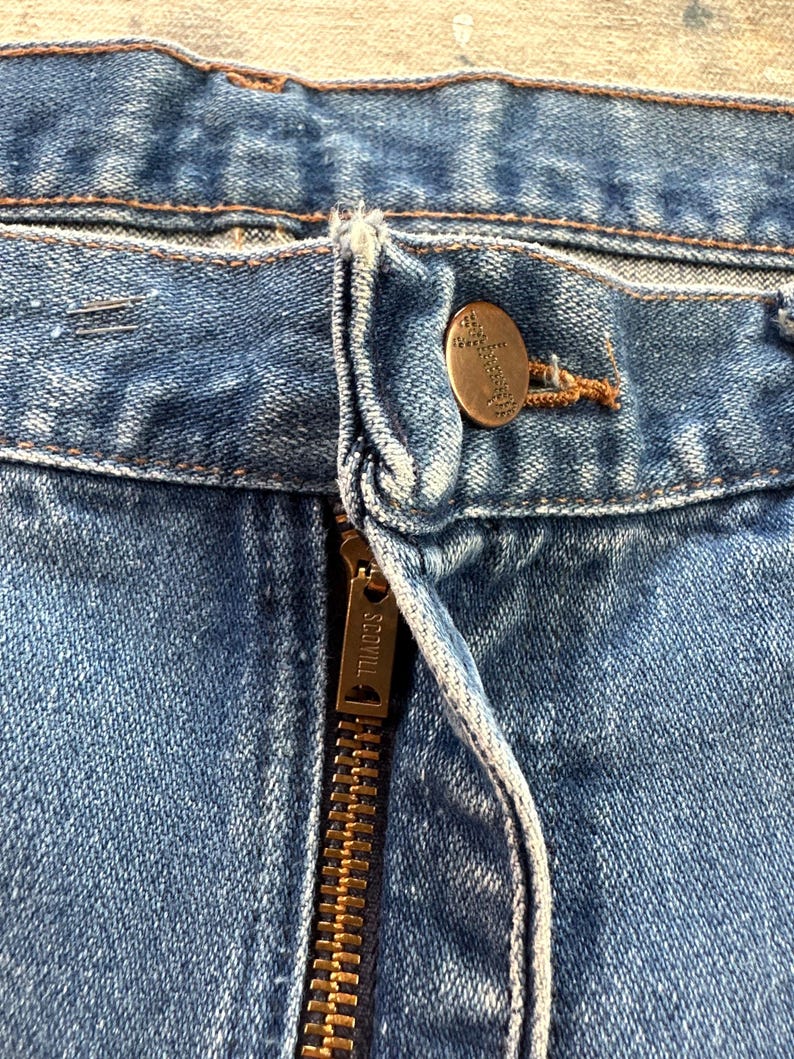 May include: Close-up of a pair of blue denim jeans. The image highlights the button and zipper area, with a brass-colored button and a zipper labeled "SCOVILL". The denim has a faded appearance.