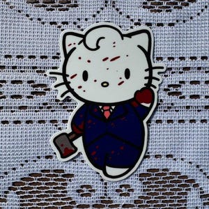 Puede incluir: Un personaje blanco de Hello Kitty con salpicaduras de sangre roja en su cara y traje. Est&aacute; sosteniendo un hacha ensangrentada en su mano derecha. El personaje lleva un traje azul oscuro con una corbata roja.