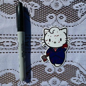 Puede incluir: Un personaje blanco de Hello Kitty con salpicaduras de sangre roja en la cara, vistiendo un traje azul y sosteniendo un hacha ensangrentada. El personaje est&aacute; en un estilo de dibujos animados.