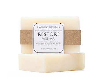 May include: Two bars of white soap with a brown paper label that reads "Rayburn's Naturals All Natural Handcrafted Locally Made Restore Face Bar Repairs Dry and Damaged Skin, Protects Against Wrinkles, Smooths Skin, Improves Skin Tone, Promotes Radiant Skin Net Wt Approx 3 oz"