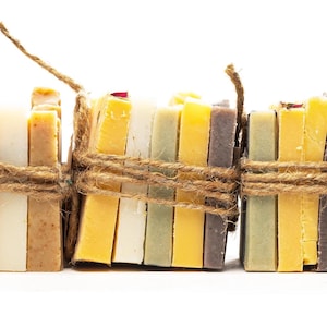 May include: Bundles of handmade soap bars in various colors, including yellow, white, and green, are tied together with twine. The soaps are arranged in neat stacks, showcasing their different textures and hues. The image is set against a clean white background.