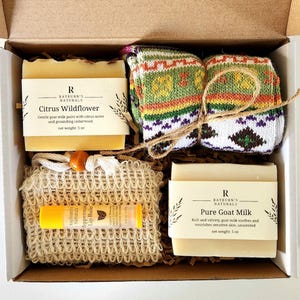 May include: A gift box containing a bar of Citrus Wildflower soap, a pair of patterned socks, a lip balm, and a bar of Pure Goat Milk soap. The soap bars are labeled with the brand "Rayburn's Naturals" and the net weight is 5 oz.