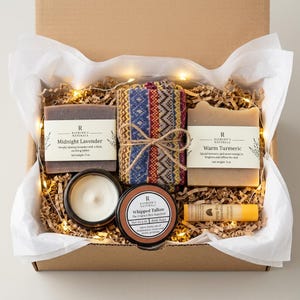 May include: A gift box containing two 5 oz soap bars labeled Midnight Lavender and Warm Turmeric, a 2 oz Whipped Tallow jar, a lip balm, and a folded patterned fabric item tied with twine, all nestled in shredded paper with string lights.