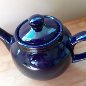 May include: A glossy, deep cobalt blue ceramic teapot with a rounded body, a curved spout, and a matching lid featuring a small knob handle. The teapot has a smooth, reflective finish and a sturdy side handle, presenting a classic, simple design.