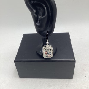 May include: A silver-toned dangle earring featuring a rectangular pendant with a beaded border. The center of the pendant contains a black background with small, colorful geometric shapes in red, blue, and white, suspended by a silver hook wire.