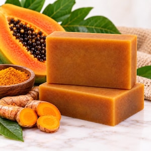 May include: Two rectangular bars of orange soap stacked on top of each other. Beside the soap are fresh papaya slices with black seeds, a small wooden bowl filled with orange turmeric powder, and several pieces of raw turmeric root.