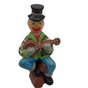 May include: A ceramic figurine of a seated clown playing a banjo. The clown wears a black top hat, a colorful plaid jacket, and blue striped pants. The banjo is brown with gray accents, and the clown is seated on a brown log.