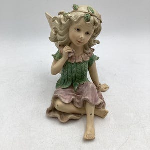 Könnte beinhalten: Eine sitzende Feenfigur mit einem ruhigen Ausdruck. Die Fee hat lockiges blondes Haar, das mit Blättern geschmückt ist, und trägt ein grünes Oberteil und einen rosa Rock. Die Figur hat zarte Flügel und besteht aus einem hellfarbigen Material.