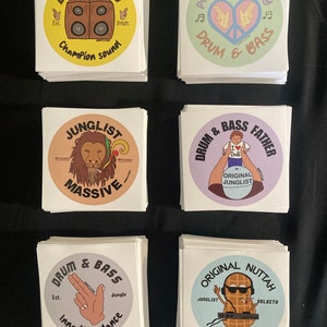 Puede incluir: Seis pilas de pegatinas redondas de colores con varios diseños relacionados con la música drum and bass. Las pegatinas presentan ilustraciones de altavoces, manos haciendo un signo de paz, un león, una persona con un corazón, un cacahuete con gafas de sol y una mano haciendo un gesto de pistola. Las pegatinas tienen texto como "Drum & Bass", "Jungle", "Wicked", "Massive", "Original Junglist", "DNB", "Peace Love Unity", "Champion Sound", "Inna the Dance", y "Original Nuttah".