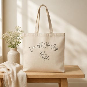 Könnte beinhalten: Beige Canvas-Tragetasche mit langen Henkeln. Auf der Tasche steht in handschriftlichem Stil der Text "Learning to Notice Joy", zusammen mit einer Stern- und Herzgrafik. Die Tasche liegt auf einem Holztisch.