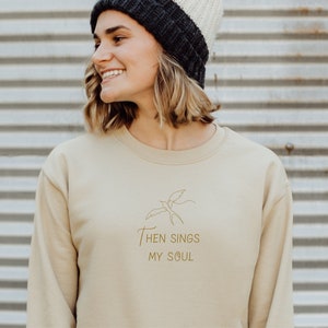 May include: A beige sweatshirt with a gold outline of a bird and the text "Then Sings My Soul".