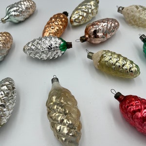 Könnte beinhalten: Eine Sammlung von 11 vintage Weihnachtsbaumschmuck aus Glas in Form von Tannenzapfen. Der Schmuck ist in verschiedenen Farben, darunter Silber, Gold, Grün, Rot und Weiß.