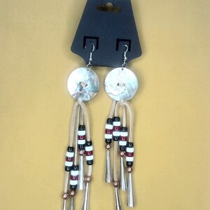 May include: A pair of silver earrings with a white and brown beaded tassel and a round, iridescent shell pendant. The earrings are hanging from a black card with the word "FRONT" printed on it.