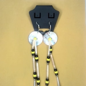 May include: A pair of earrings with a white shell disc, bone beads, and black and yellow beads hanging from each. The earrings are displayed on a black stand with the word "FRONT" on a white card above.