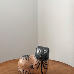 Op de afbeelding: Een keramieken kattenbeeldje met een zwarte kop en lichaamsmarkeringen. De kat heeft grote, ronde ogen en is geschilderd in een bruin en zwart kleurenschema. Het beeldje staat op een houten oppervlak.