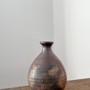 May include: A small, round, dark brown ceramic vase with a narrow neck and a flared rim. The surface features a textured, mottled finish with patches of lighter tan and black tones, showing a rustic, uneven appearance across the rounded body.