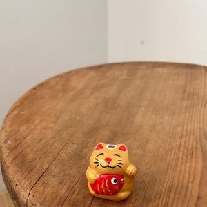 May include: A small, yellow ceramic Maneki-neko cat figurine with red detailing. The cat holds a red fish and has a smiling face. The figurine is on a round, wooden surface.