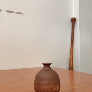 Może przedstawiać: Mały, brązowy ceramiczny wazon o zaokrąglonym korpusie i wąskiej szyi stoi na drewnianym stole. Wazon ma teksturowaną powierzchnię i ciemnobrązowy kolor. Drewniany kij baseballowy opiera się o ścianę w tle.