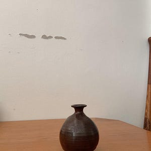 以下が含まれることがあります： 丸みを帯びたボディと細い首を持つ、小さな茶色の陶器の花瓶。花瓶は木製のテーブルの上に置かれています。背景はプレーンなオフホワイトの壁です。