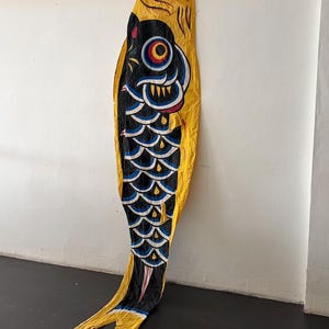 May include: A long, fabric carp-shaped windsock featuring a vibrant yellow body with a black, white, and blue scale pattern. The fish has a prominent eye with red, yellow, and blue rings, a red mouth opening, and a bifurcated tail at the bottom.