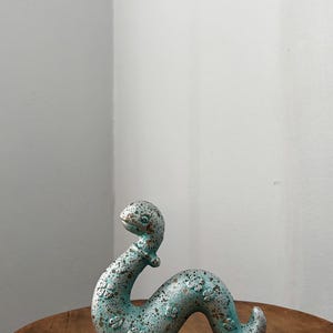 Puede incluir: Figura de serpiente de cerámica con acabado moteado turquesa y plateado. La escultura tiene un cuerpo curvado y una cabeza levantada, con pequeños detalles florales. La figura está colocada sobre una superficie de madera.
