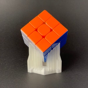 May include: A solved Rubik's Cube with orange, blue, and white sides, displayed on a white 3D-printed stand. The cube is a classic puzzle toy, showcasing a combination of colors and geometric design.