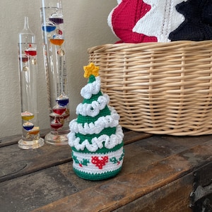 May include: A small crocheted Christmas tree ornament with green and white yarn, a red heart, and a yellow star on top. The ornament is sitting on a wooden surface.