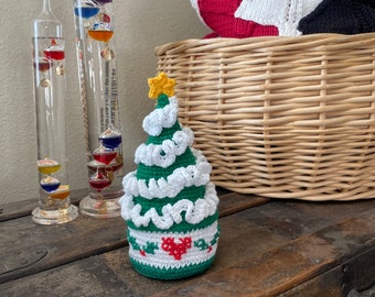 May include: A small crocheted Christmas tree ornament with green and white yarn, a red heart, and a yellow star on top. The ornament is sitting on a wooden surface.
