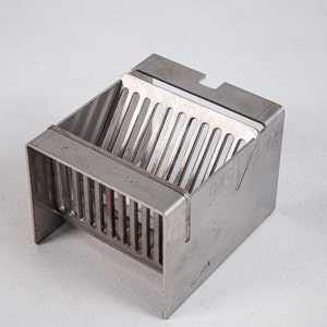 May include: A silver metal grate with a rectangular shape and a series of parallel bars. The grate is designed to fit into a larger structure, possibly a drain or ventilation system.