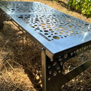 Puede incluir: Una mesa plegable de plata con un diseño único de patrón circular. La mesa está hecha de metal y tiene un logotipo negro que dice "Campfire" en la superficie.
