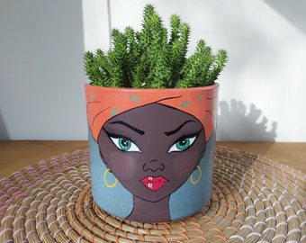 May include: A ceramic planter with a painted design of a woman's face with green eyes and red lips. The woman is wearing a bright orange headscarf with a pattern. The planter is filled with a green succulent plant.