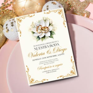 Puede incluir: Invitaci&oacute;n de boda marfil con detalles dorados y dise&ntilde;o floral. La invitaci&oacute;n presenta el texto "Nuestra Boda" y los nombres "Valeria & Diego". Se muestran anillos de boda dorados. La invitaci&oacute;n est&aacute; rodeada de adornos dorados.
