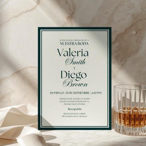 May include: Wedding invitation with a dark green border and elegant script. The invitation includes the names Valeria Smith and Diego Brown, along with the date and time of the ceremony. A glass of amber liquid sits to the right.