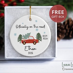 May include: A round white ceramic Christmas ornament featuring a red truck carrying a green pine tree, blue snowflakes, and the text "Officially on the road!", "BEEP! BEEP!", "Ethan", and "2026". The ornament hangs by a gold string inside a white box.