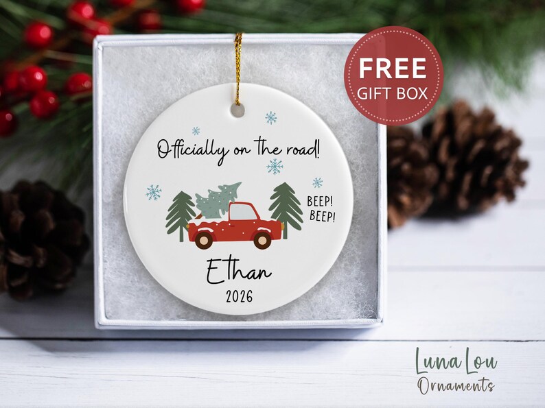 May include: A round white ceramic Christmas ornament featuring a red truck carrying a green pine tree, blue snowflakes, and the text "Officially on the road!", "BEEP! BEEP!", "Ethan", and "2026". The ornament hangs by a gold string inside a white box.