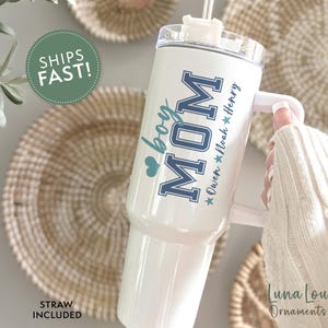 May include: White stainless steel tumbler with a blue and white design that says "Mom" with a heart and "boy" underneath. The tumbler has a handle and a straw. The text below the design says "Owen * Noah * Henry"