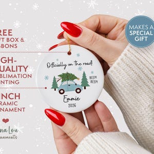May include: A round white 7.62 cm ceramic ornament featuring a light blue car carrying a green Christmas tree on its roof. The text reads Officially on the road!, BEEP! BEEP!, Emmie, and 2026. The design includes small blue snowflakes and green pine trees.