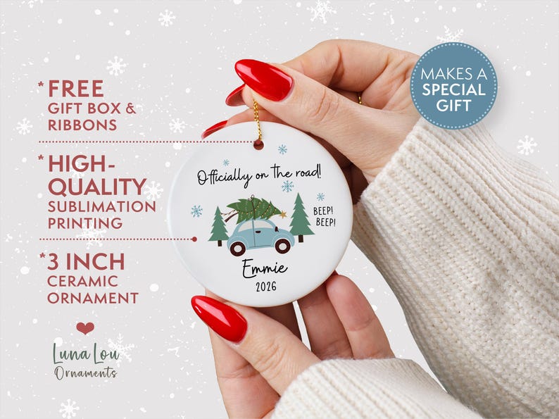 May include: A round white 7.62 cm ceramic ornament featuring a light blue car carrying a green Christmas tree on its roof. The text reads Officially on the road!, BEEP! BEEP!, Emmie, and 2026. The design includes small blue snowflakes and green pine trees.