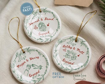 May include: Three round white ceramic ornaments with gold hanging strings. Each features a green wreath design, red text, and illustrations of engagement rings, hands, or champagne glasses with dates like 11.11.2026 and 10.17.26.
